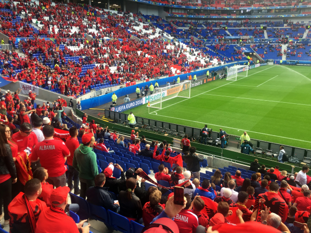 Ein volles Stadion mit Zuschauern, einige sitzen und andere stehen, die ein Fußballspiel beobachten, mit Bannern, einem Torpfosten und Deckenleuchten.
