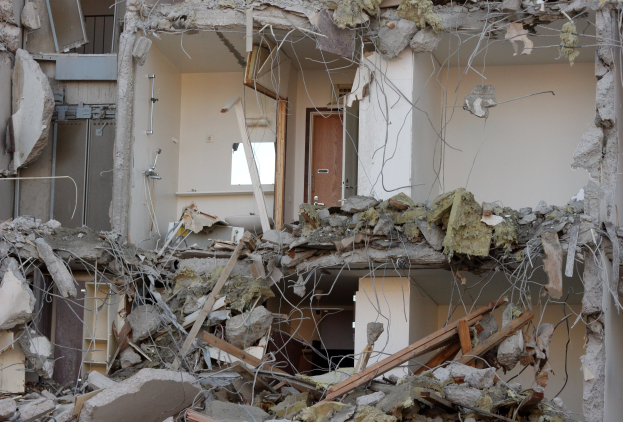 Zusammengebrochenes Gebäude mit verstreuter Trümmern, Schutt, gebrochenem Holz und einer beschädigten Wand mit einer Tür im Hintergrund.