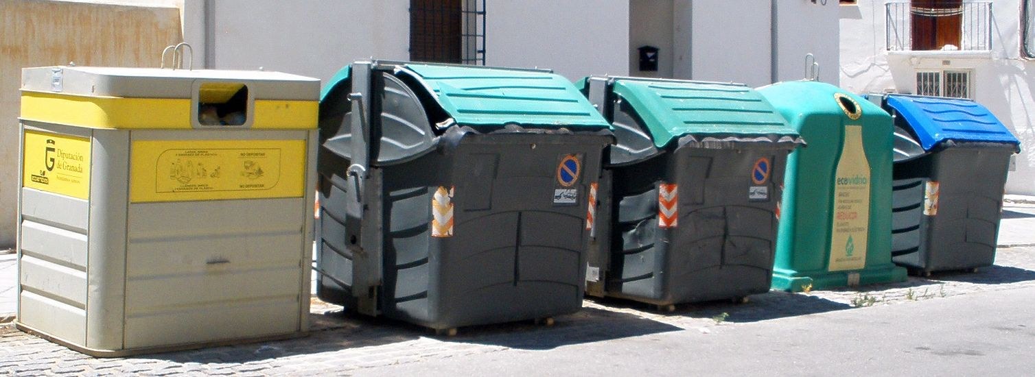 Eine Reihe bunter Mülltonnen, die auf einer Straße neben einem Gebäude mit Fenstern, Geländern und einer Tür aufgereiht sind.