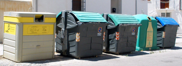 Eine Reihe bunter Mülltonnen, die auf einer Straße neben einem Gebäude mit Fenstern, Geländern und einer Tür aufgereiht sind.