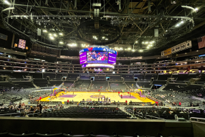 Eine große Arena voller Zuschauer, die ein Basketballspiel im Staples Center verfolgen, mit einem großen Bildschirm, einer Flagge, Anzeigetafeln und einer von Lampen geschmückten Decke.