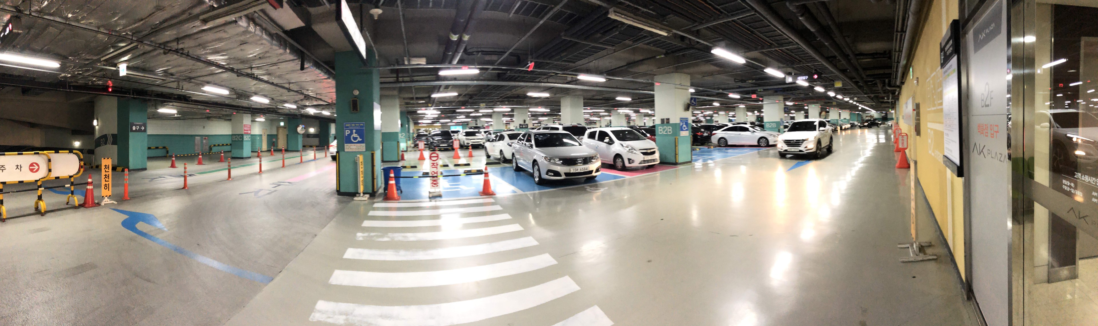 Ein mehrgeschossiges Parkhaus mit zahlreichen geparkten Autos, Deckenleuchten, Verkehrskegeln, Schildern und Stützpfeilern.