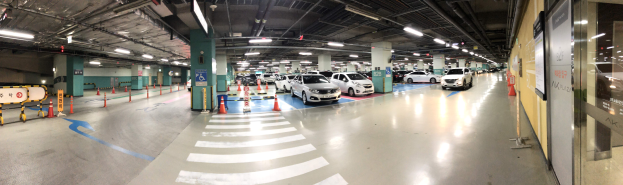 Ein mehrgeschossiges Parkhaus mit zahlreichen geparkten Autos, Deckenleuchten, Verkehrskegeln, Schildern und Stützpfeilern.