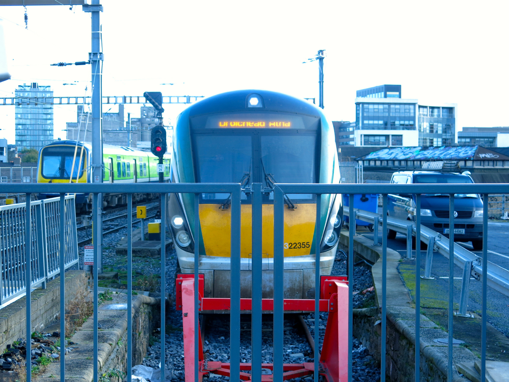 Zwei Züge auf dem Gleis mit einem Zaun im Vordergrund, Masten auf dem Land, einer mit einer Signalleuchte, ein Auto auf der Straße rechts, Gebäude im Hintergrund und der Himmel oben.