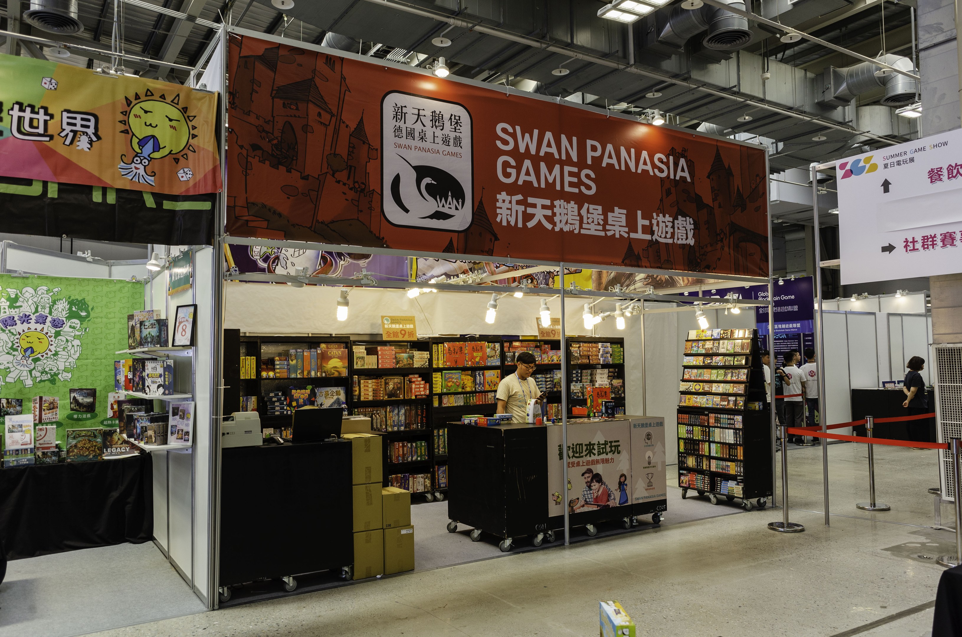 Messestände für "Swan Panasia Games" mit Bücherregalen, einem Menschen an einem Tisch mit einem Laptop, Kartons, Bändern, Bannern, Deckenleuchten und Menschen im Hintergrund.
