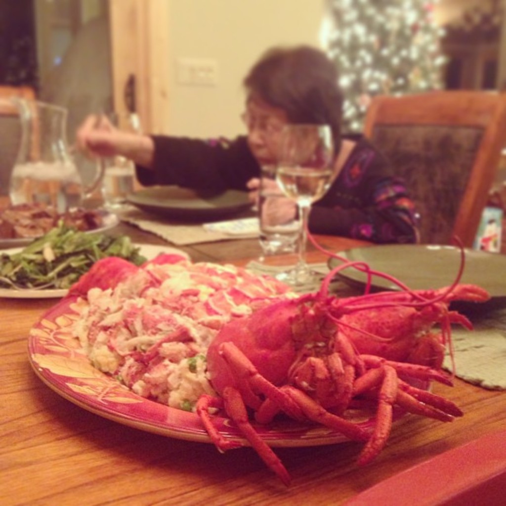 Eine Frau sitzt an einem Tisch und hält Chopsticks in der Hand, mit verschiedenen Lebensmitteln, einem Wasserglas und einem Krug vor sich; ein Weihnachtsbaum ist oben rechts zu sehen, sowie eine Tür mit Steckdosen im Hintergrund.