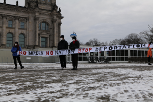 Eine Gruppe von Menschen steht im Schnee und hält eine Fahne mit der Aufschrift "No Border, No Nation, Stop Deportation". Im Hintergrund sind Fahrräder, Pflanzen, Bäume und ein Zaun zu sehen.