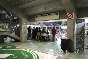 Gruppe von Menschen vor einem sitzenden Publikum in einem Stadion mit Schildern, die "Boston Celtics vs New York Jets" lesen, einem Müllcontainer, einer Tafel mit Text, einer Treppe mit Geländern und einem Dach mit Deckenlampen.
