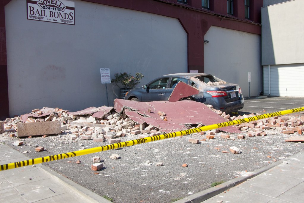 Eine zerbrochene Wand mit verstreuten roten Ziegelsteinen, ein geparktes Auto, eine Topfpflanze und ein Gebäude mit einer 'Kautionsgeschäfte'-Reklame auf einem Schild.