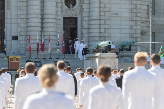 Gruppe von Menschen in weißen Marineuniformen, die auf einer Treppe vor einem Gebäude mit Säulen, einem Podium und Fahnen während einer Abschlussfeier stehen.