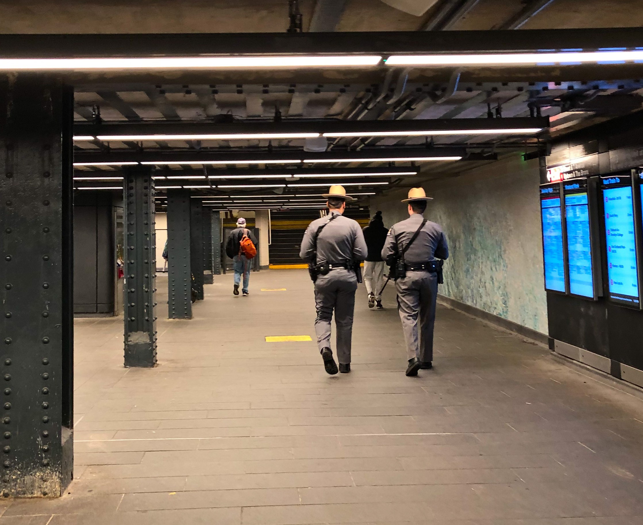 Zwei Polizeibeamte in Uniform gehen durch eine U-Bahn-Station, mit Wandbildschirmen auf der rechten Seite, Säulen und Deckenleuchten im Hintergrund.