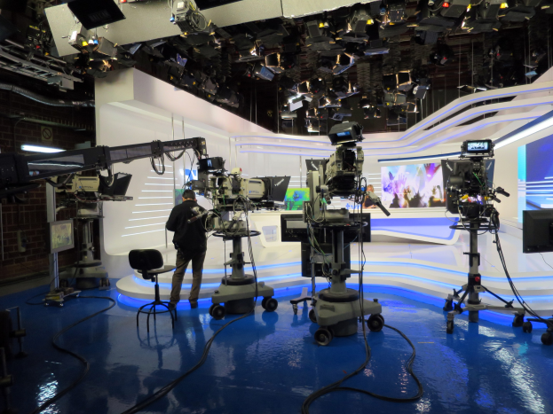 Ein Mann in einer Live-Broadcast-Umgebung hält ein Mikrofon, während er vor einem Fernseher steht, umgeben von Kameras, einem Stuhl, einem Anzeigebildschirm und Deckenleuchten.