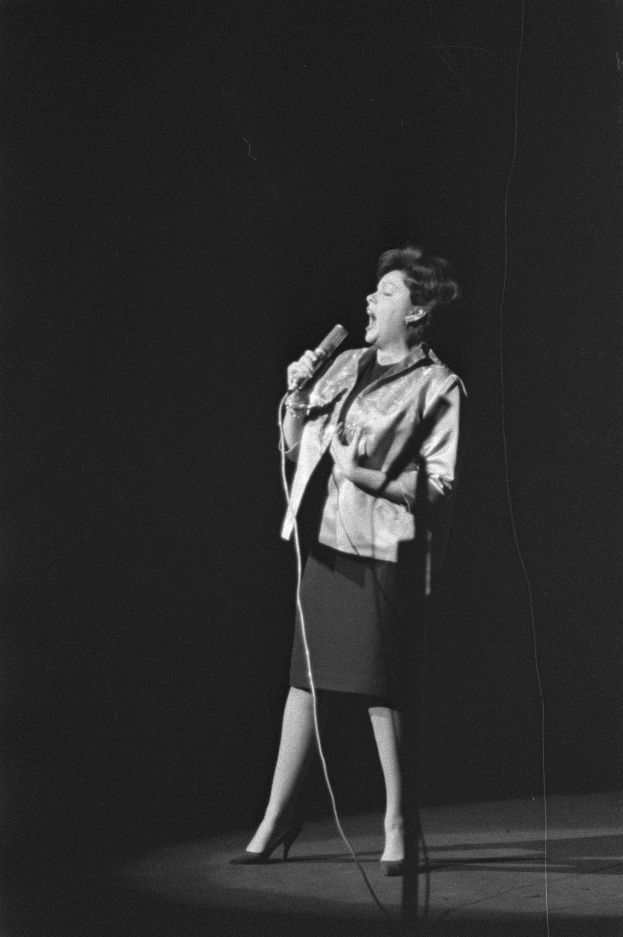 Eine Frau in einem schwarzen und weißen Outfit aus den 1950er-Jahren singt fröhlich auf einer von einem Scheinwerfer beleuchteten Bühne in ein Mikrofon, ihr Haar ist im klassischen 50er-Jahre-Stil frisiert.