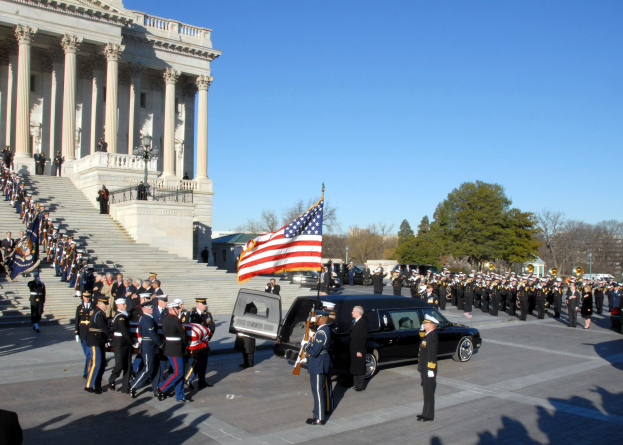 Trauerprozession mit Menschen in Mützen und Fahnen vor dem U.S. Capitol Building, Fahrzeuge auf der Straße, Treppen mit Geländern, Laternenpfähle, Bäume und ein Himmel im Hintergrund.