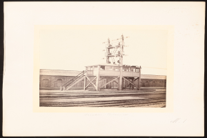 Ein Schwarz-Weiß-Foto eines Bahnhofs mit einer zentralen Signalbox, umgeben von Bahnschienen und einem Gebäude im Hintergrund, mit Text am unteren Bildrand.