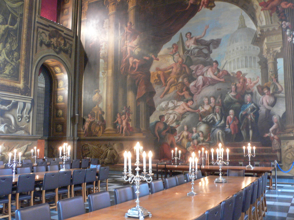 Ein Raum mit Tischen und Stühlen, jeder Tisch hat Kerzenständer und im Hintergrund eine Wand mit einem Bogen und Gemälden.