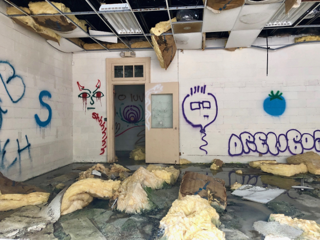 Ein Raum mit Graffiti an den Wänden, eine Tür im Hintergrund, Deckenstangen und Lampen sowie verschiedene Gegenstände, die auf dem Boden verstreut sind und tragische Ereignisse in der Detroit-Region symbolisieren.