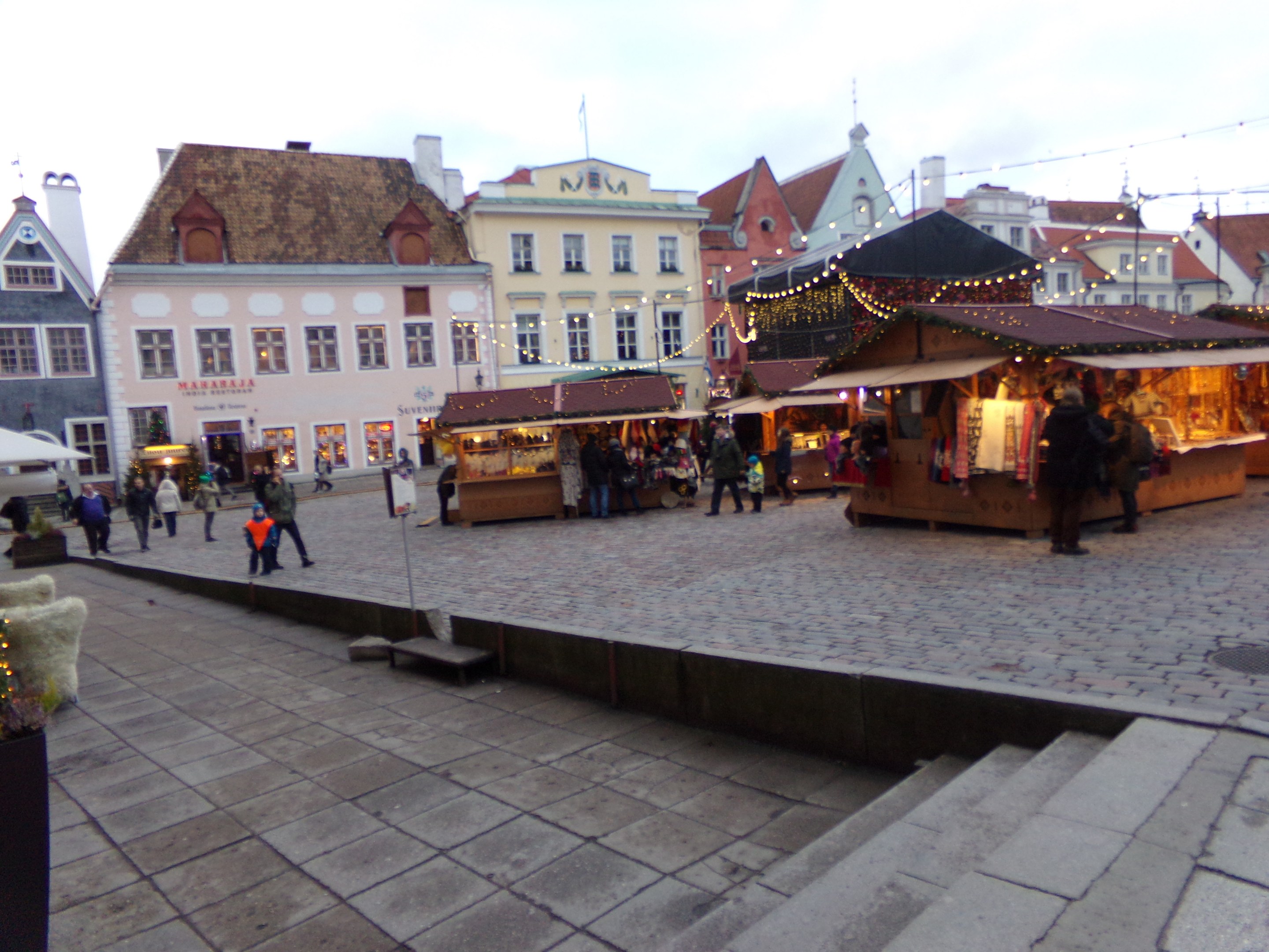 Ein belebter Weihnachtsmarkt in Tallinn, Estland mit Menschen um geschmützte Stände, festlicher Beleuchtung, bewölktem Himmel, Treppen führfürend und Topfpflanzen im Bereich.