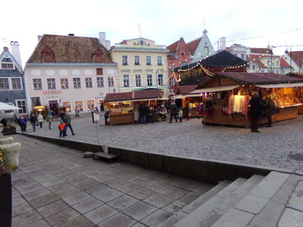 Ein belebter Weihnachtsmarkt in Tallinn, Estland mit Menschen um geschmützte Stände, festlicher Beleuchtung, bewölktem Himmel, Treppen führfürend und Topfpflanzen im Bereich.