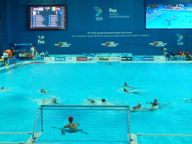 Gruppe von Athleten beim Wasserballspielen in einem Pool während der FINA-Weltmeisterschaften 2016 in Kazan, mit Zuschauern auf der linken Seite und einem Anzeigebrett mit Event-Logo im Hintergrund.