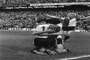 Drei Fussballspieler feiern ein Tor auf dem Boden, mit jubelnder Menge und schwenkenden Fahnen im Hintergrund, dargestellt in Schwarz-Weiss.