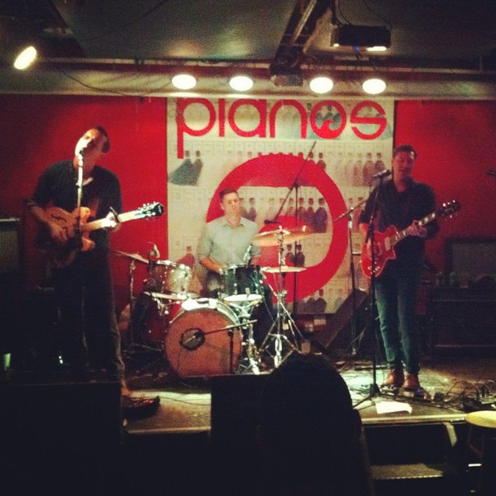 Drei Männer auf der Bühne: einer spielt Schlagzeug in der Mitte, und zwei spielen Gitarre und singen links und rechts daneben, mit einem Banner an der Wand und Lampen an der Decke.