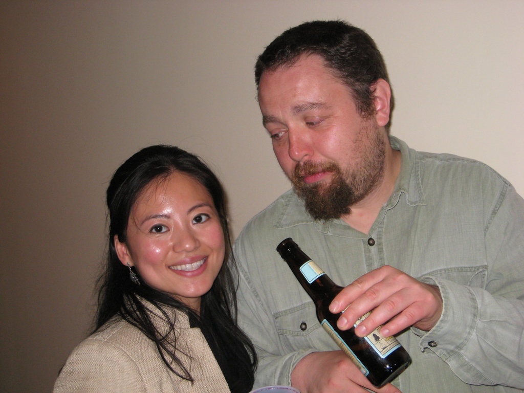 Ein Mann h├Ąlt eine Flasche, waehrend er eine l├Ąchelnde Frau ansieht, beide stehen vor einer Wand.