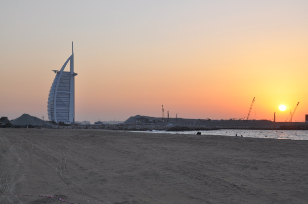 Das Burj Al Arab Hotel in Dubai, VAE, ragt prominent gegen einen Sonnenuntergang mit Sand, Wasser, Kränen, Bäumen und einem klaren blauen Himmel.