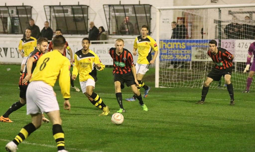 Ein Sportfeld mit mehreren Männern, die Fußball spielen, einem Netz auf der rechten Seite, einer Tafel mit Text im Hintergrund und Zuschauern, die hinter der Tafel stehen, sowie einer Wand und Fenstern weiter im Hintergrund.
