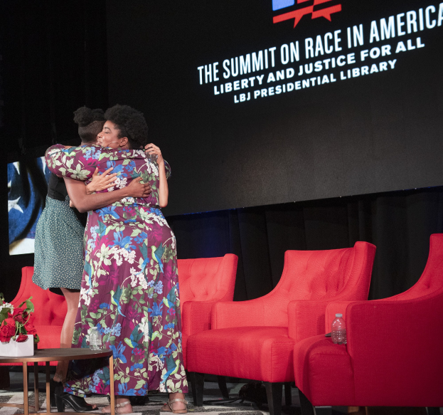 Zwei Frauen, die als L.B.J. Präsidentin und First Lady Michelle Obama identifiziert wurden, umarmen sich auf einer Bühne vor einem Bildschirm mit der Aufschrift "The Summit on Race in America", mit einem Tisch und Stühlen auf beiden Seiten.