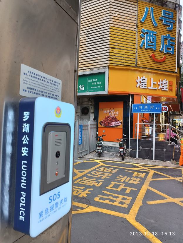 Straßenszene in Hong Kong mit Gebäuden, Schildern, Motorrädern, Fußgängern, Verkehrskegeln, Bäumen und einem sichtbaren "SOS"-Notrufsignal an einer Wand.