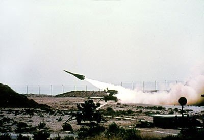 Eine Rakete wird von einem Militärfahrzeug in einer Wüstenlandschaft gestartet, mit Vegetation und Bodenobjekten, gegen eine Umzäunung im Hintergrund unter einem Himmel mit Text oben.