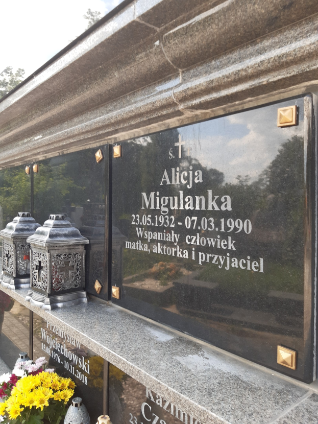 Denkmalstein mit einer Plakette mit der Aufschrift "St. Alicia Migulanka" vor einem Gebäude, umgeben von Blumenvasen und anderen Gegenständen, mit Bäumen und einem klaren blauen Himmel im Hintergrund.