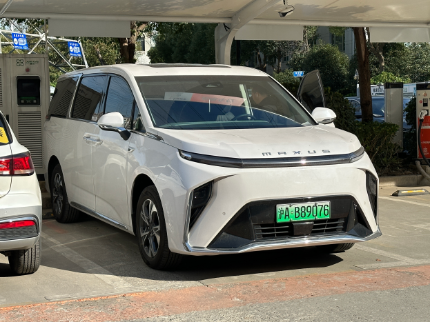Zwei Autos, ein Toyota Mirai und ein Lexus, nebeneinander auf einem Parkplatz mit einer Zapfsäule auf der rechten Seite, Bäume und Gebäude im Hintergrund.
