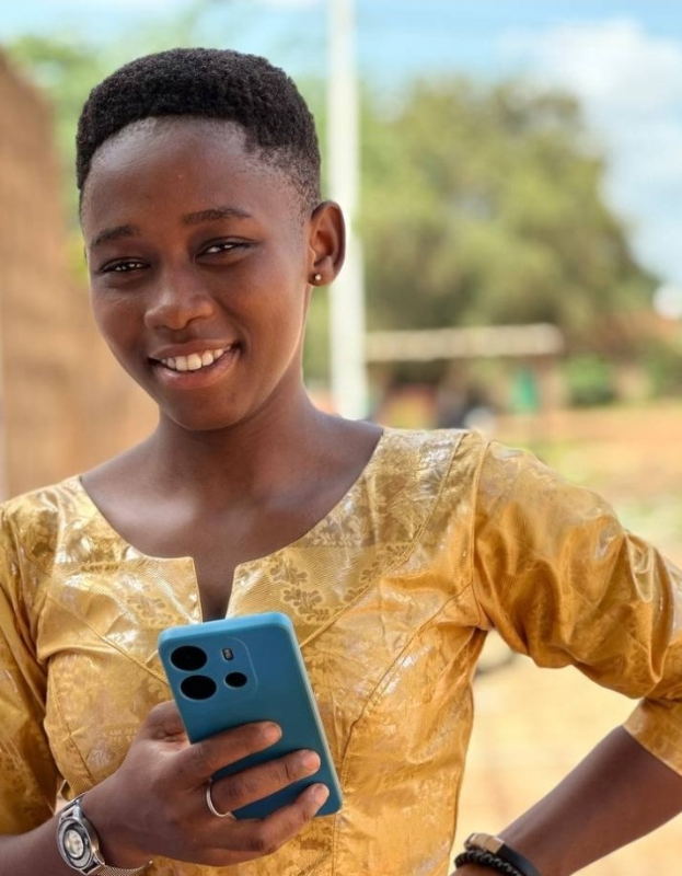 Ein junges Mädchen in einem gelben Kleid hält ein blaues Telefon und lächelt vor einer Wand, einem Pfahl, Bäumen und Himmel.