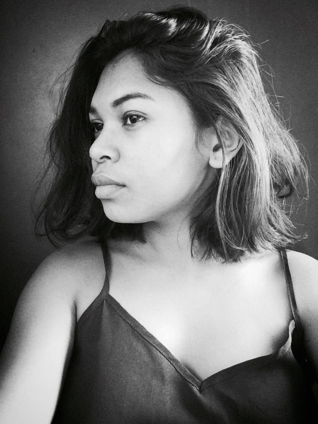 Eine selbstbewusste Frau in einem schwarzen Kleid posiert in einem Schwarz-Weiß-Foto, steht mit selbstbewusster Haltung vor einer Wand, ihr glatter Bob und das natürliche Make-up unterstreichen den selbstbewussten Ausdruck.