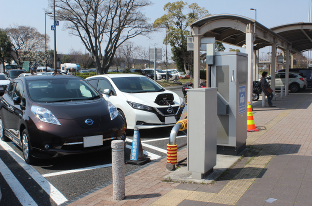Elektroauto-Ladestation in Japan mit Autos auf der Straße, Verkehrskegeln, einer Person auf dem Gehweg, einem Schuppen, Polen, Lampen, Schildern, Bäumen, Pflanzen und einem Himmel im Hintergrund.