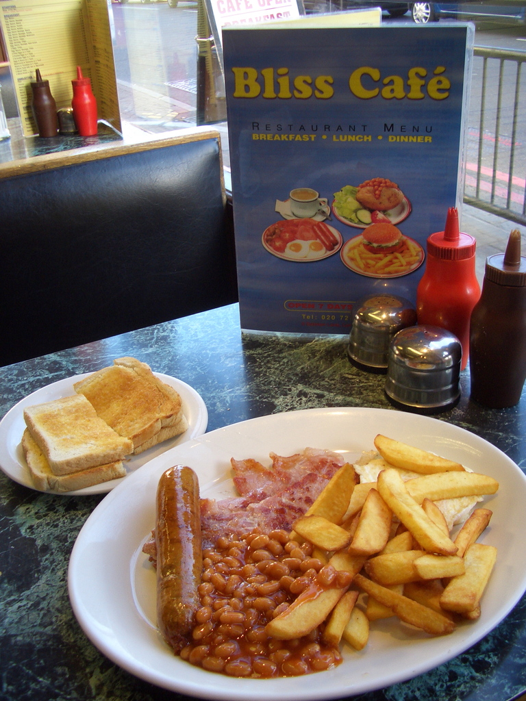Ein Teller mit Lebensmitteln auf einem Tisch, eine Speisekarte mit der Aufschrift "Bliss Cafe", Salz- und Pfefferstreuer, Soßen- und Ketchupflaschen, Banner auf dem Boden und ein Fahrzeugrad im Hintergrund.