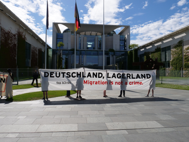 Eine Gruppe von Menschen mit einem Transparent mit der Aufschrift "Deutschland, Lagerland Migration ist kein Verbrechen" vor einem Fenster mit Fenstern, umgeben von Gras, einem Metallzaun, Pflanzen, B├Ąumen und Fahnenmasten unter einem bew├Âlktem Himmel.