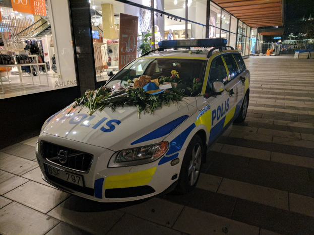Ein mit Blumen und Blättern geschmückter Polizeiwagen steht vor einem Geschäft mit Glaswänden in der Nacht, das Schaufensterpuppen in Kleidern und beleuchtete Schilder zeigt.