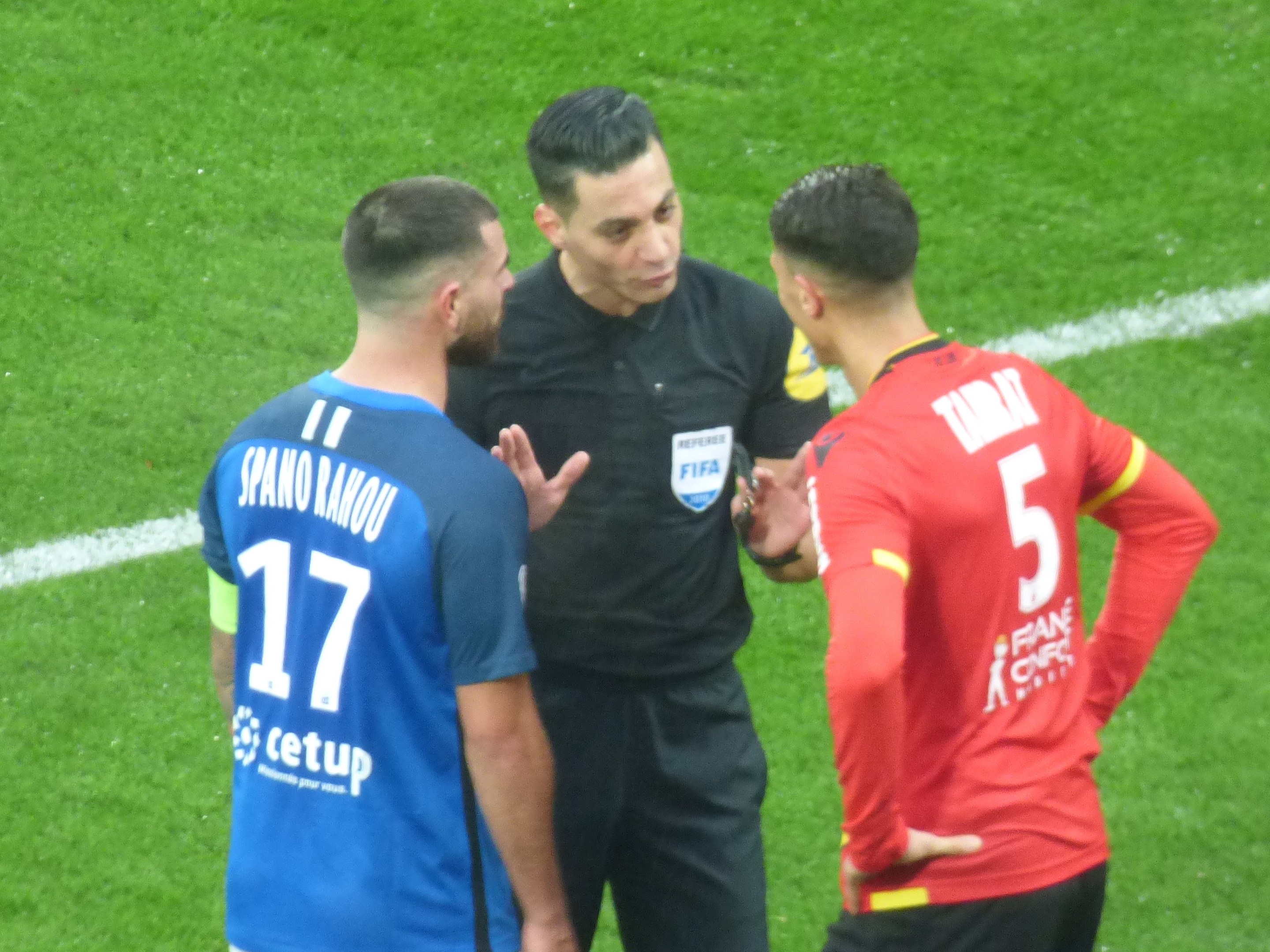 Schiedsrichter spricht mit zwei Männern auf einem Fußballfeld, einer trägt eine schwarze Hose und ein schwarzes Hemd, der andere ein blau-rotes T-Shirt mit Text und Brille.