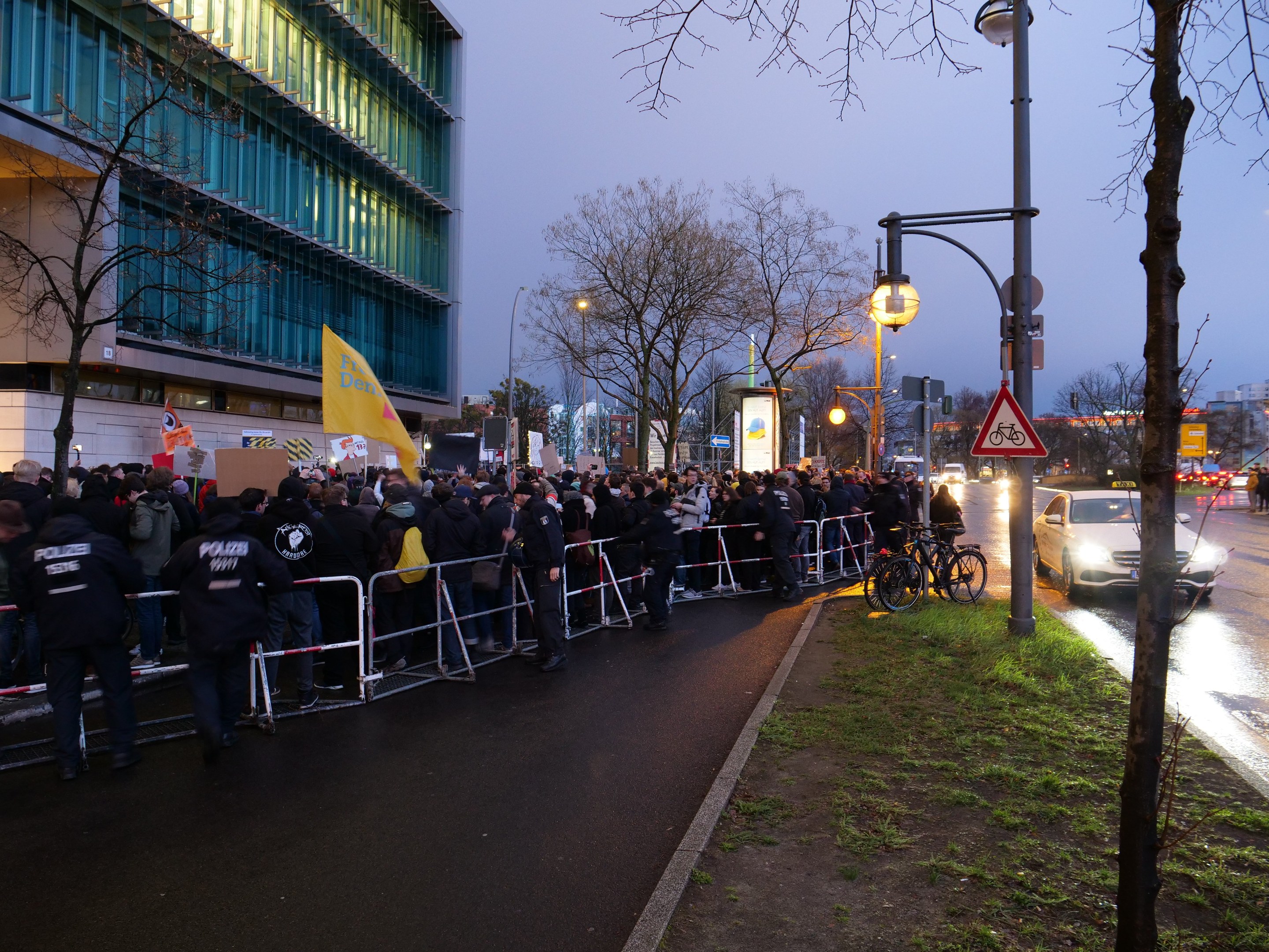 Eine große Gruppe von Menschen mit Schildern steht vor einem Gebäude mit Barrikaden, Fahrrädern, Laternen, Schildern, Bäumen und Gras unter einem sichtbaren Himmel, was auf eine Protestaktion in Berlin hindeutet.