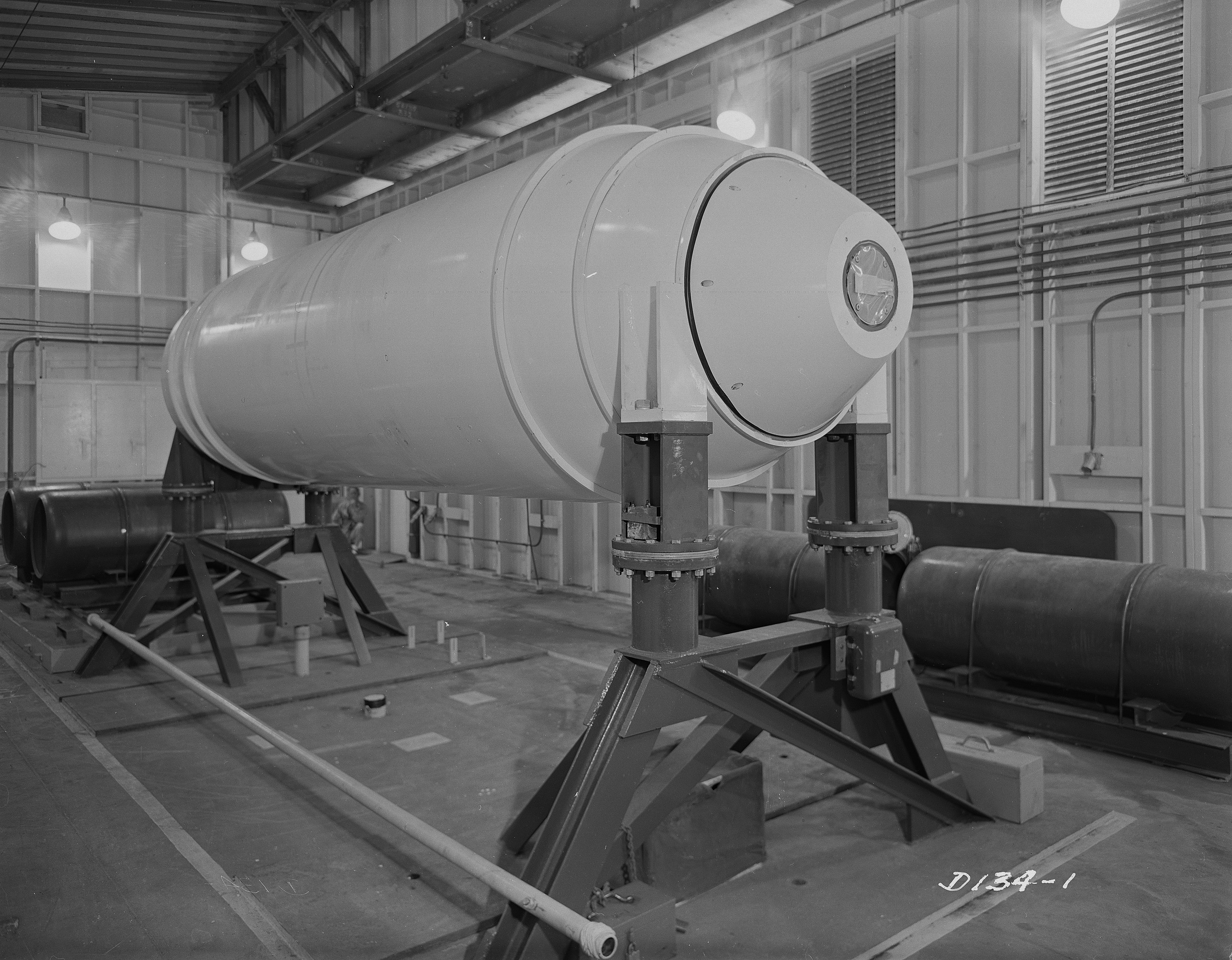 Große zylindrische Wasserstoffbombe auf einem Ständer in einer industriellen Fabrikumgebung mit Rohren, Lampen und einer Wand im Hintergrund.