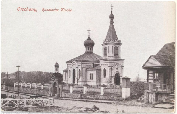 Schwarz-weiß-Fotografie einer russisch-orthodoxen Kirche in Olschany, Russland, umgeben von Häusern, Bäumen, Pfählen und einem Zaun unter einem klaren Himmel, mit Text oben.