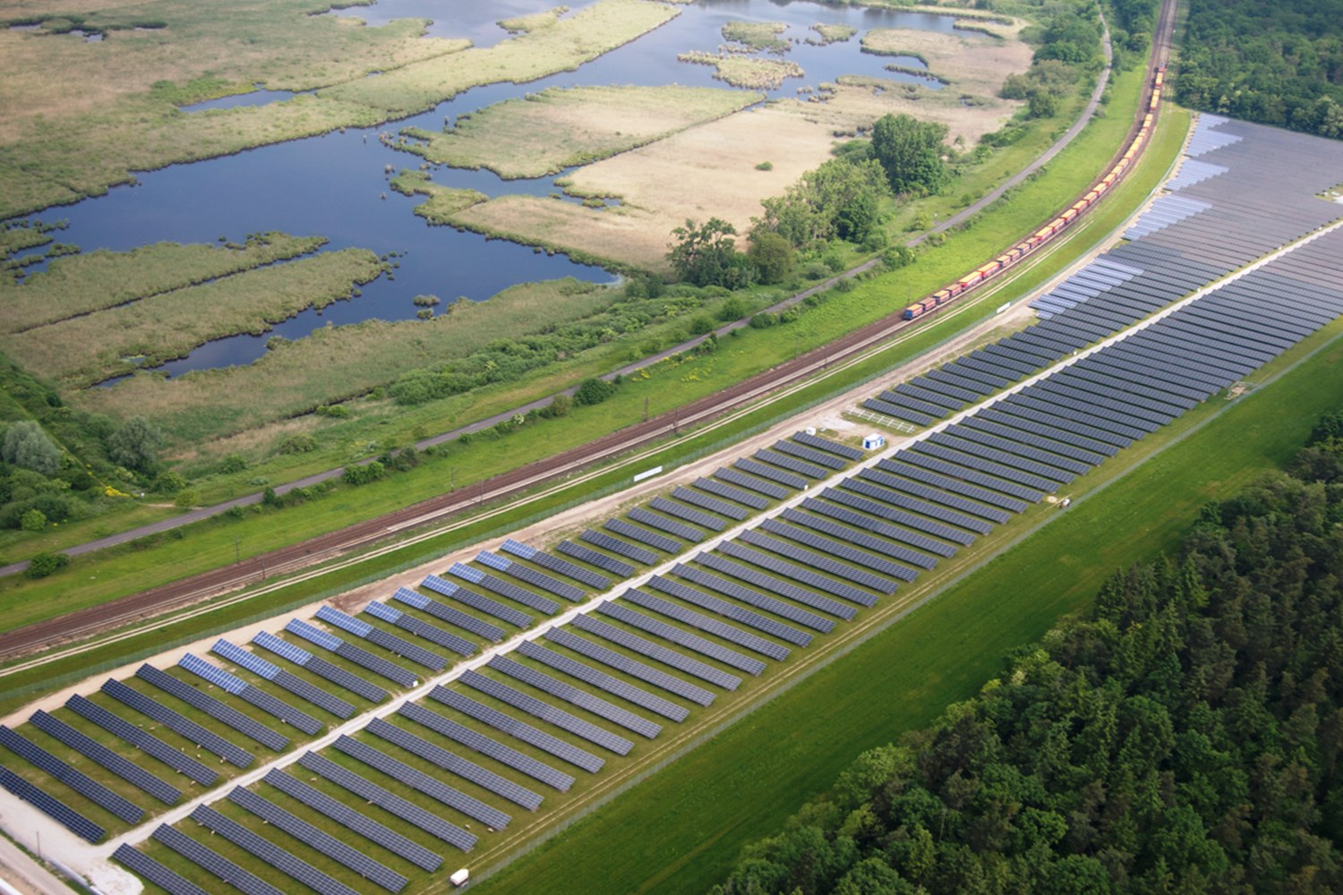 Luftaufnahme eines großen Solarparks mit Solarpanelen auf einem Feld, umgeben von Bäumen, Gras, Pflanzen und Wasser, mit einem Zug auf einer nahen Bahnstrecke.