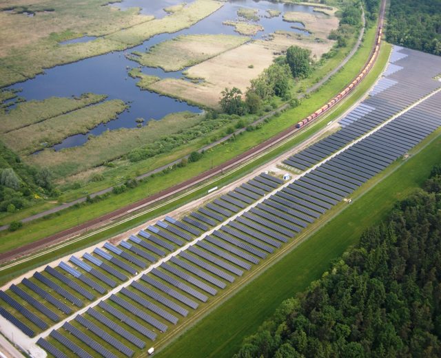 Luftaufnahme eines großen Solarparks mit Solarpanelen auf einem Feld, umgeben von Bäumen, Gras, Pflanzen und Wasser, mit einem Zug auf einer nahen Bahnstrecke.