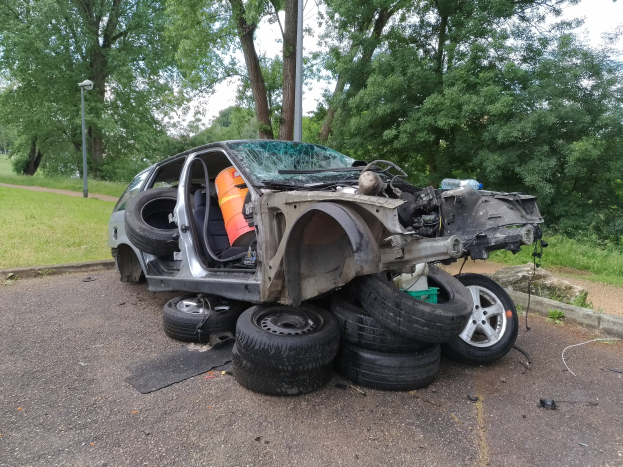 Ein umgestürzter Wagen am Straßenrand umgeben von Vegetation und Trümmern unter einem bewölkten Himmel.