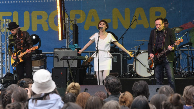 Vier Personen auf der Bühne: eine Frau in einem weißen Kleid singt in der Mitte, flankiert von zwei Männern, die Gitarren spielen, mit einer Menge unten und Lautsprechern an der Wand im Hintergrund.