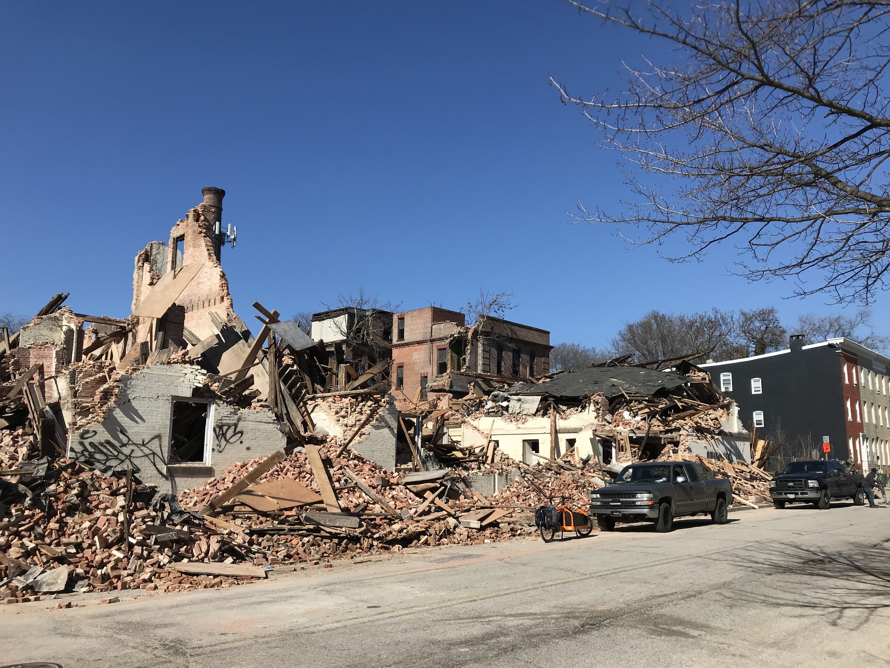 Eingestürztes Gebäude an einer Straßenecke mit verstreuter Trümmer, Fahrzeugen, Passanten, Bäumen und einem klaren Himmel.