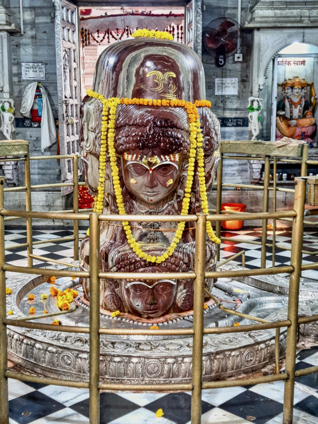 Eine steinerne Statue des Herrn Shiva in einem Hindu-Tempel, geschmückt mit Girlanden und von einem Geländer umgeben, mit zusätzlichen Statuen, einem Ventilator und einer Wand im Hintergrund.